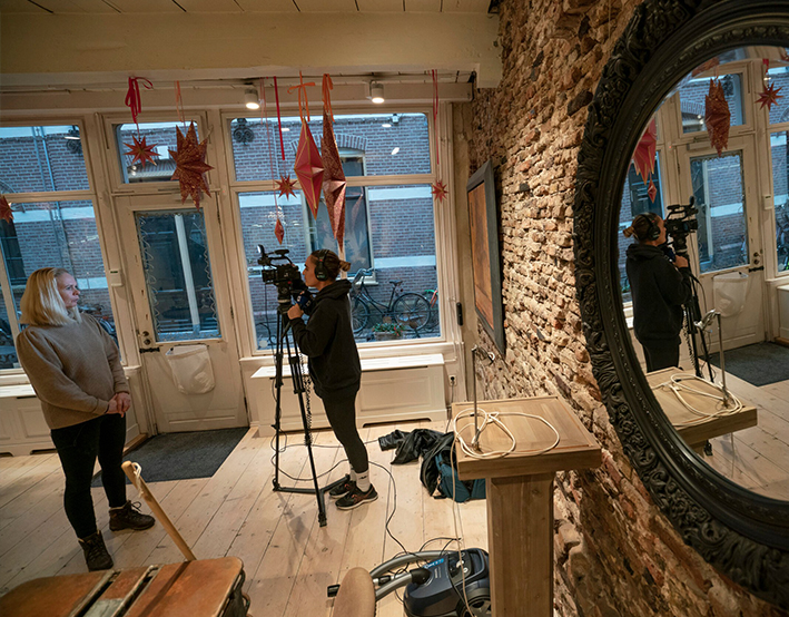 Een cameraman filmt een vrouw in een kamer met houten vloeren en bakstenen muren. Een grote spiegel reflecteert de scène. Decoratieve sterren hangen aan het plafond en fietsen zijn zichtbaar door de ramen.