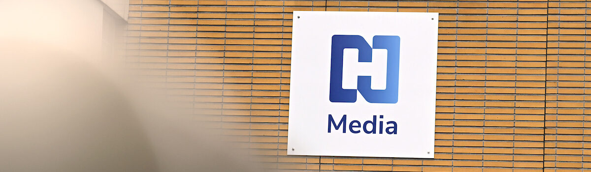 Op een bord aan de bakstenen muur staat het blauw-witte logo met de tekst 'Media', een verwijzing naar het uitgebreide Jaarverslagen NH Media.