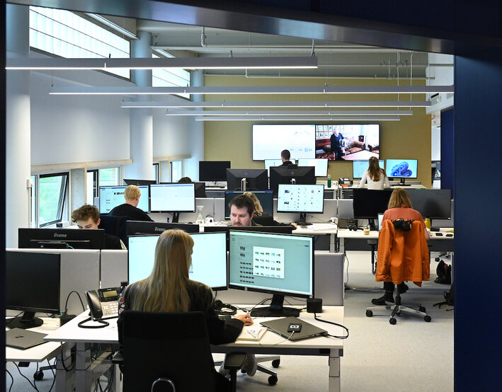 Een moderne kantoorruimte die bruist van activiteit, terwijl mensen zich concentreren op taken aan bureaus die zijn uitgerust met meerdere monitoren. Grote ramen verlichten de kamer waar een scherm inhoud weergeeft en hints geeft naar verleidelijke vacatures, wat de levendige sfeer versterkt.