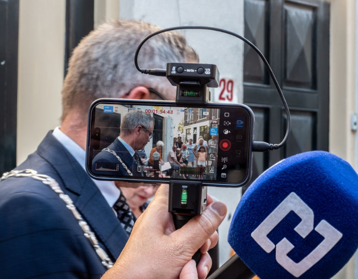 Een persoon houdt een smartphone vast waarop een man in pak met ketting wordt opgenomen, gedeeltelijk weggedraaid, terwijl een andere microfoon met een blauw hoesje zichtbaar is op de voorgrond. Op het scherm van de telefoon zijn verschillende mensen en gebouwen te zien.